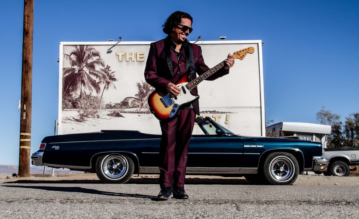 Male musician portrait Johnny San Francisco wearing red suit playing sunburst electric guitar with black convertible behind parked under white billboard