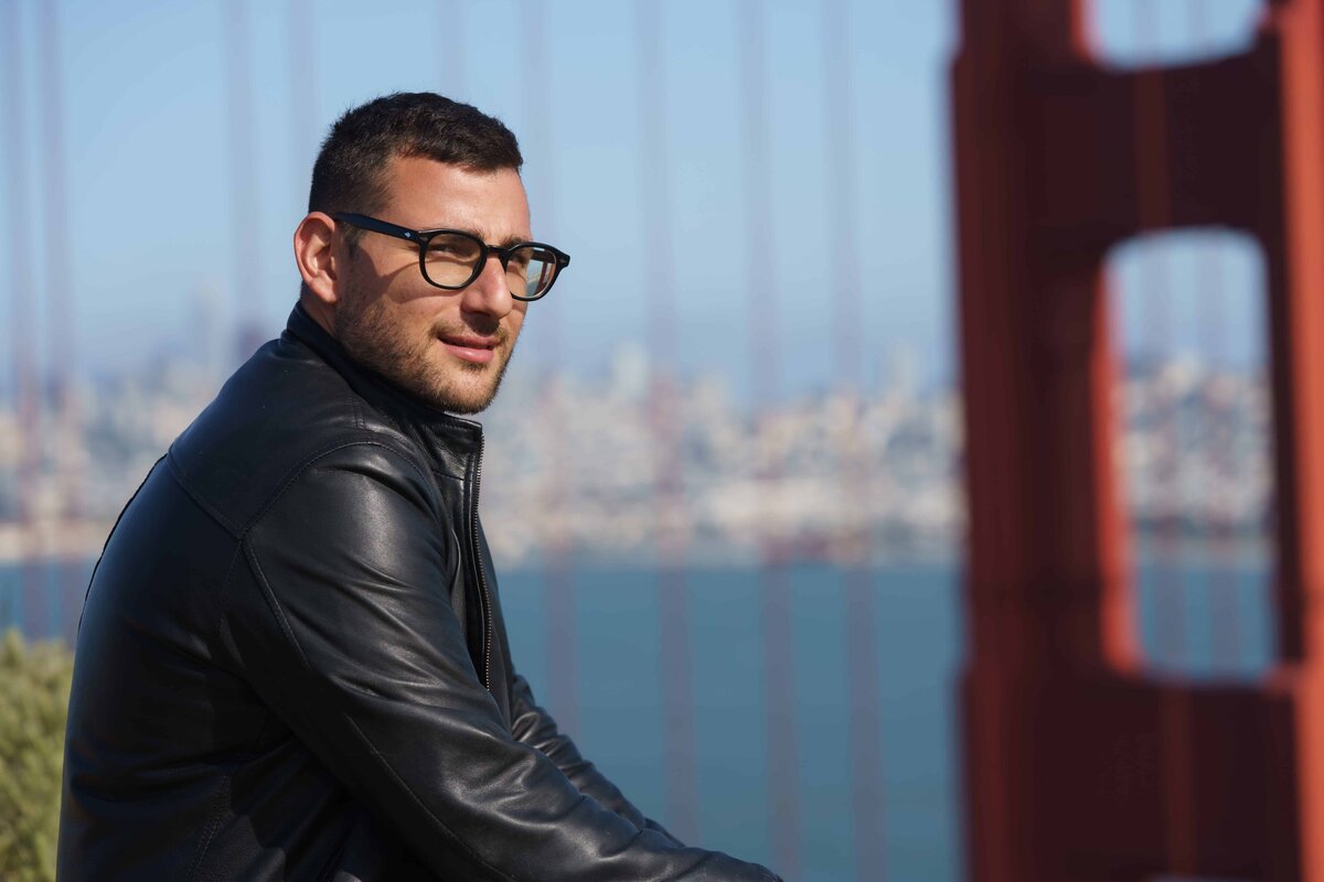 Man wearing a black leather jacket and glasses looking over his shoulder with the golden gate bridge and the city of san francisco in the background