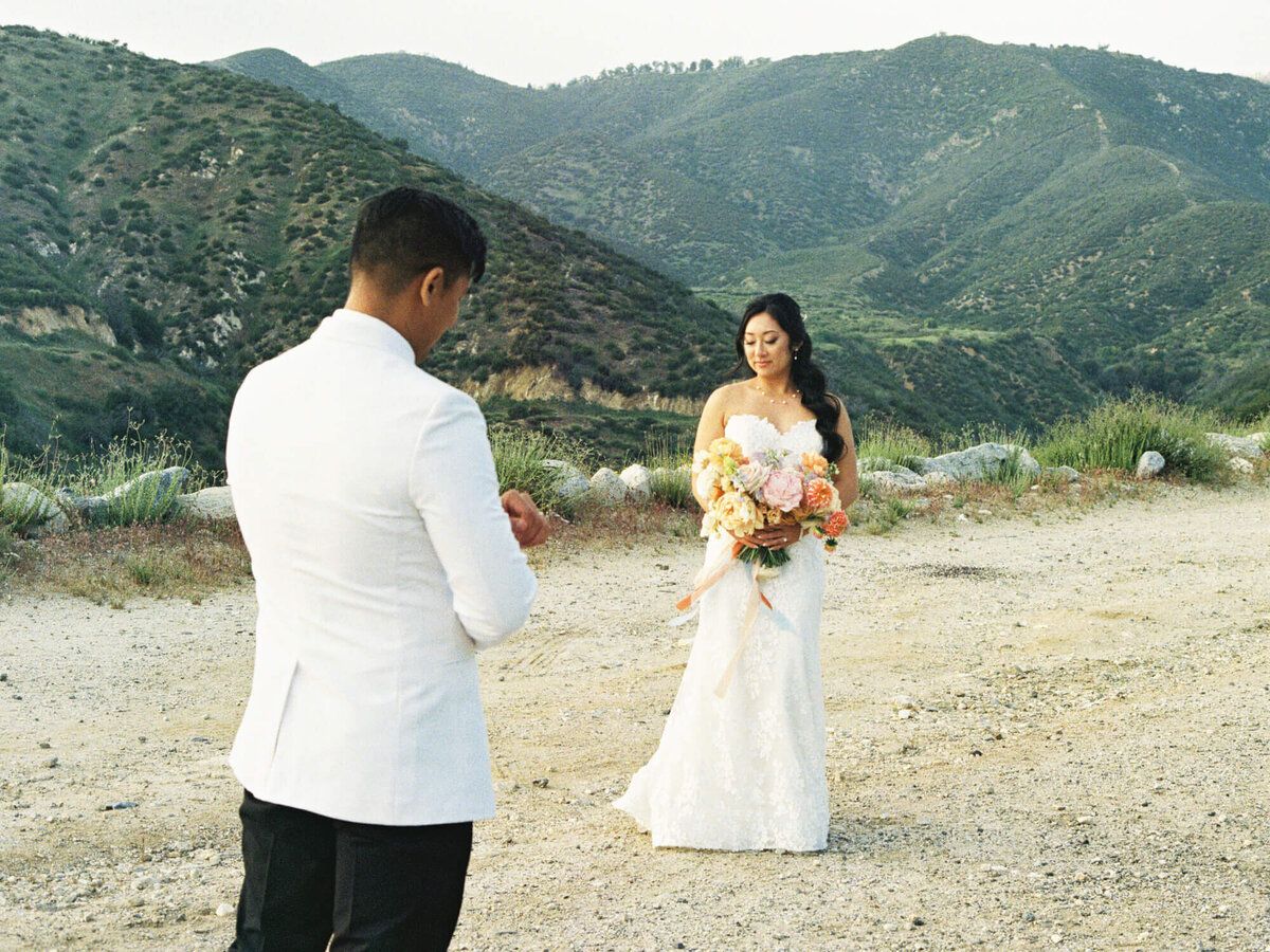 Bride holding a bouquet, standing on a mountain path. A groom in a white jacket faces her. The mood is serene and romantic, with lush hills as a backdrop.
