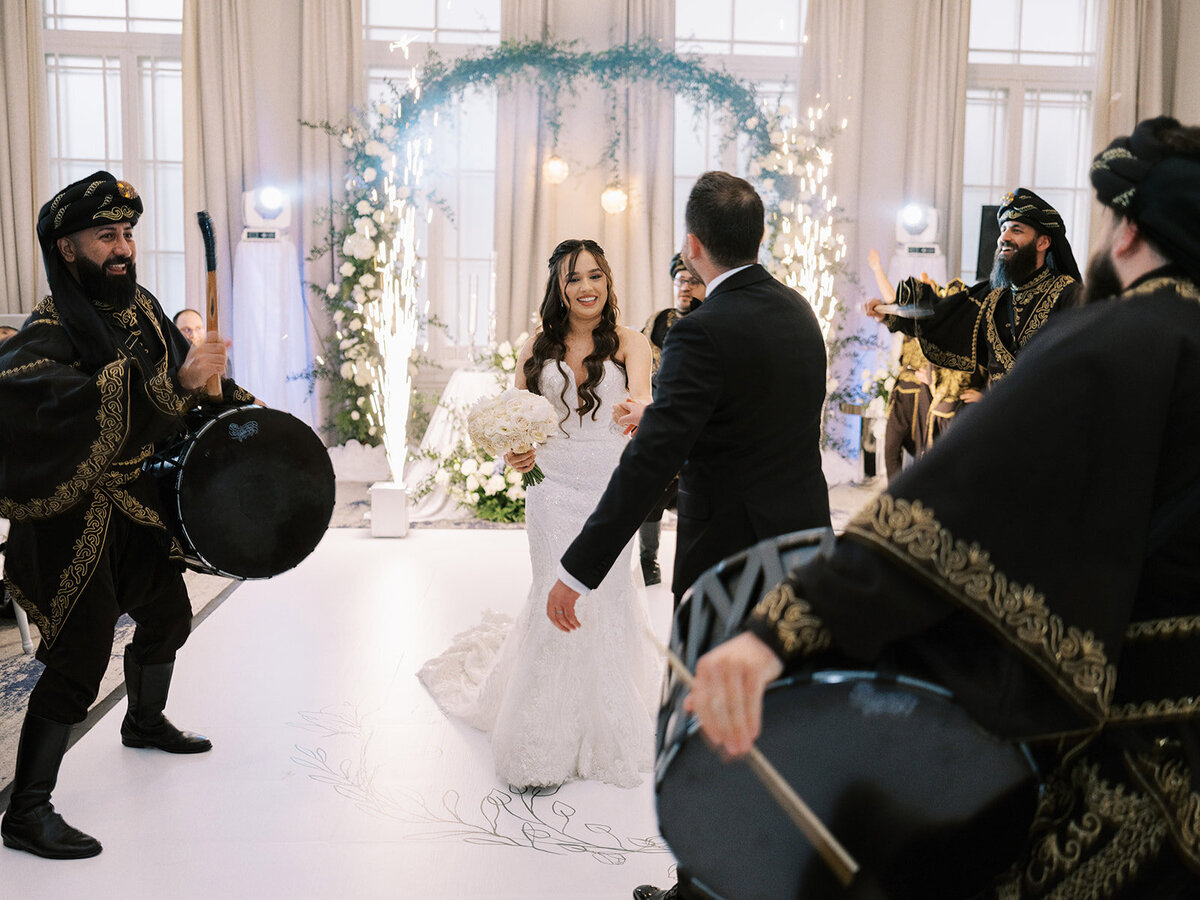 A bride and groom dance together at their classic Calgary wedding, surrounded by four musicians dressed in traditional attire, playing large drums. The venue is elegantly decorated with flowers and lights, capturing the essence of a timeless celebration at the Fairmont Palliser Wedding in Calgary.