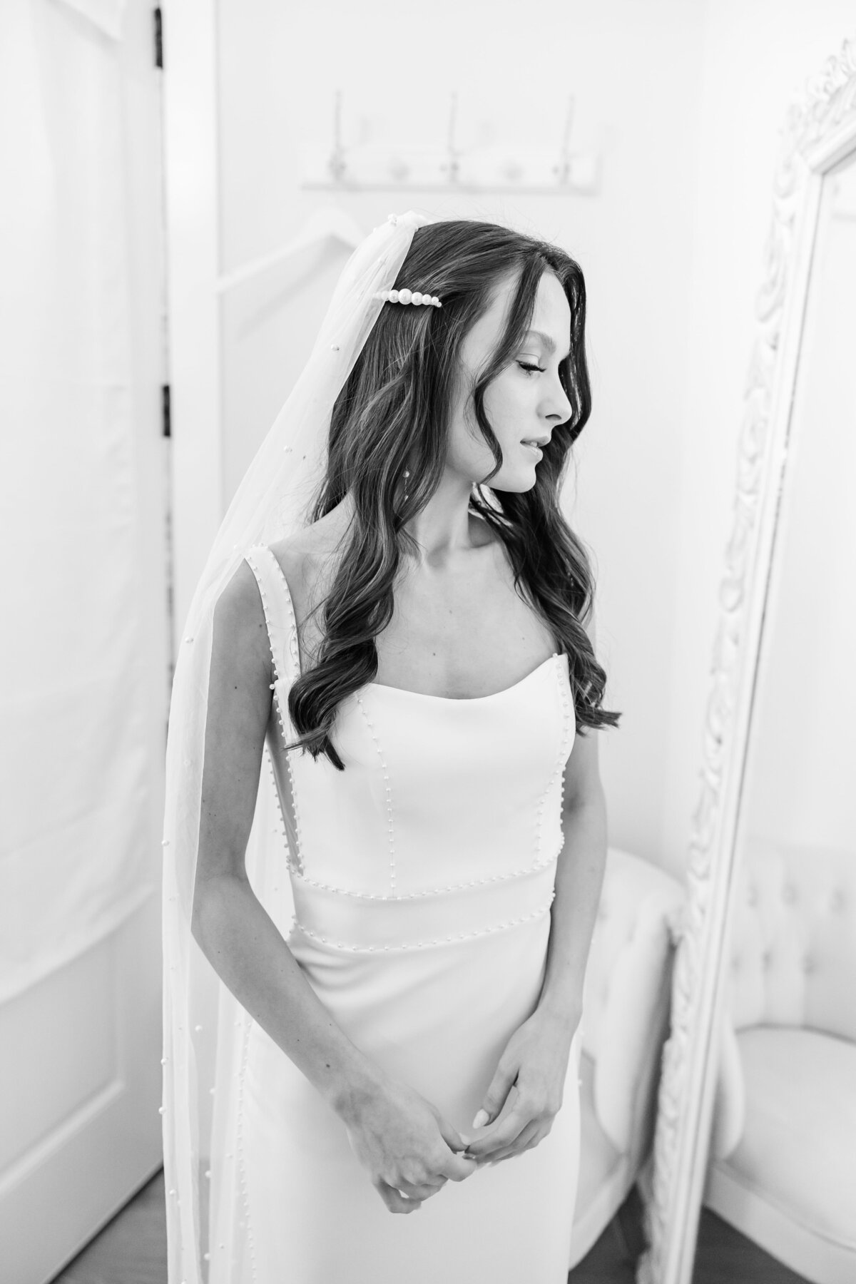 A black and white image of a bride staring at her wedding dress while in front of a mirror.