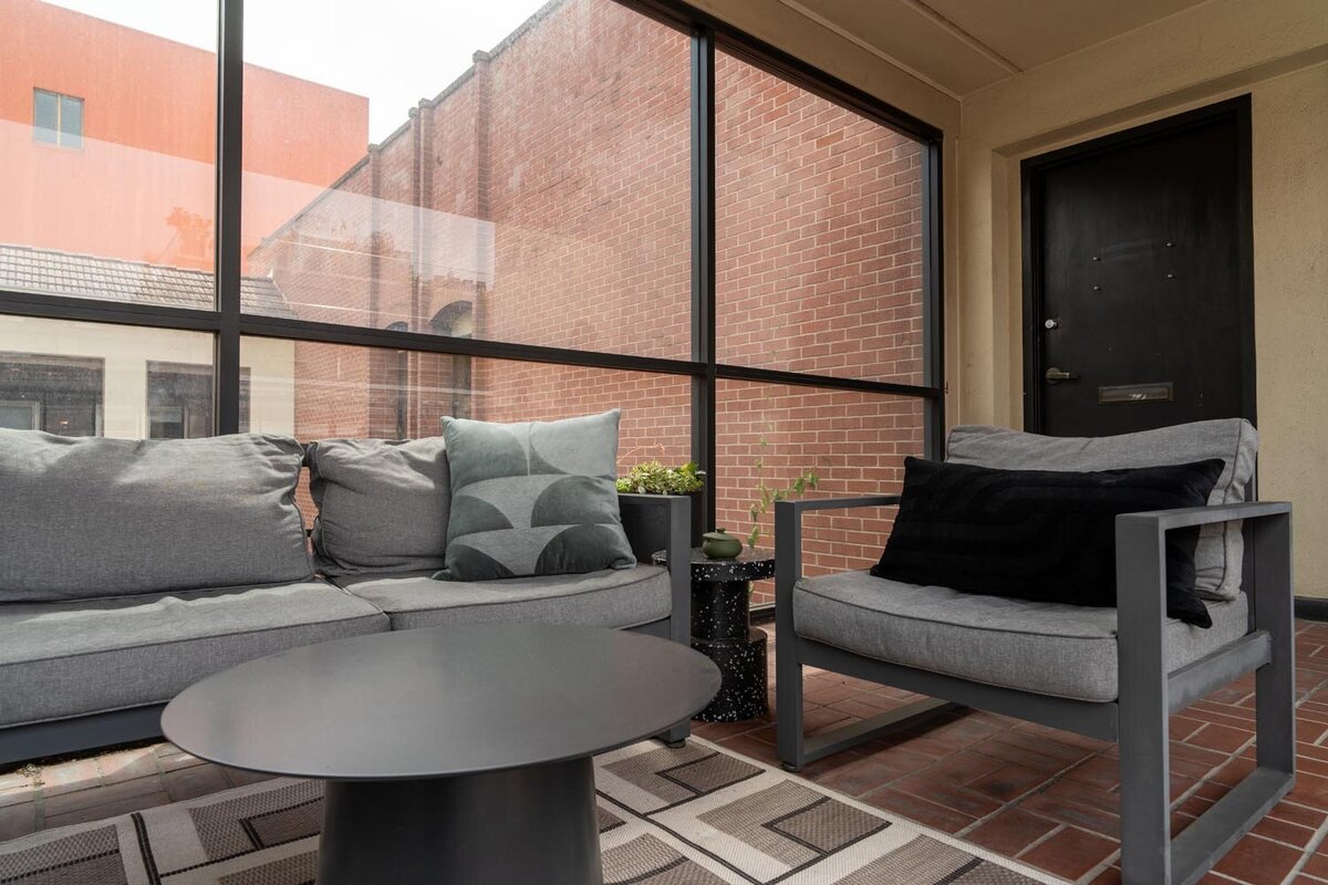 Patio area with gray cushioned lounge seating, a black metal coffee table, and views of adjacent brick buildings through floor-to-ceiling windows.