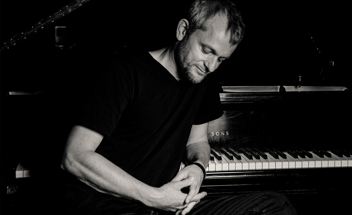 Classical musician portrait black and white Patrick Kirst leaning against grand piano keys looking down wearing black t shirt