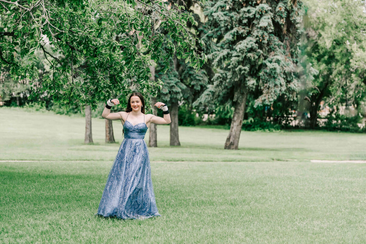 a brunette wearing a purple dress standing in grass with trees behind her is flexing her muscles.