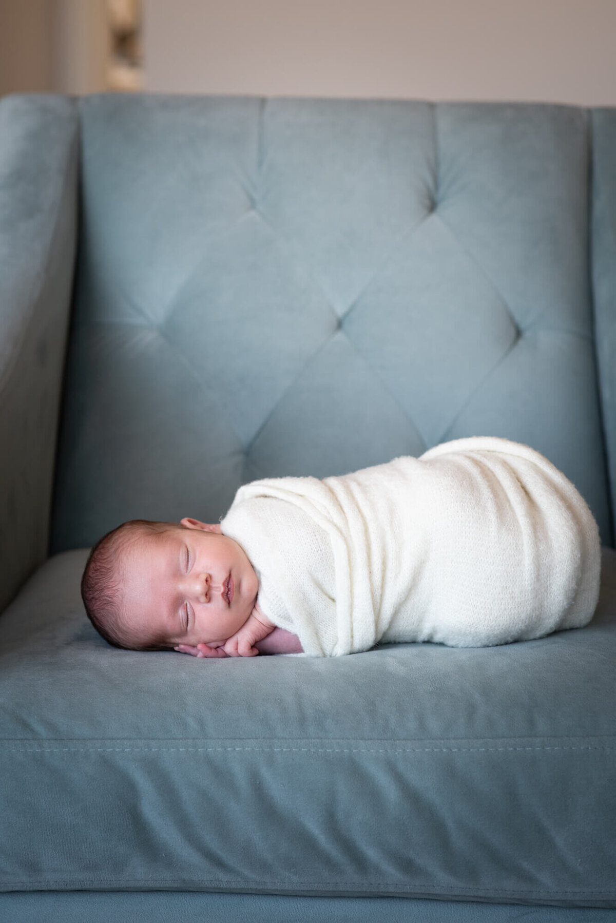 An image from a Denver Newborn Photographer of a newborn baby sleeping in a blue chair while wrapped in a white swaddle