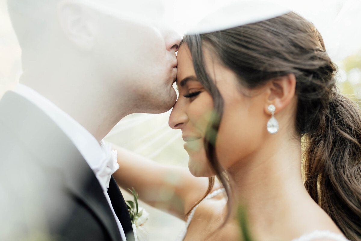 Groom kisses bride's forehead under veil. 
