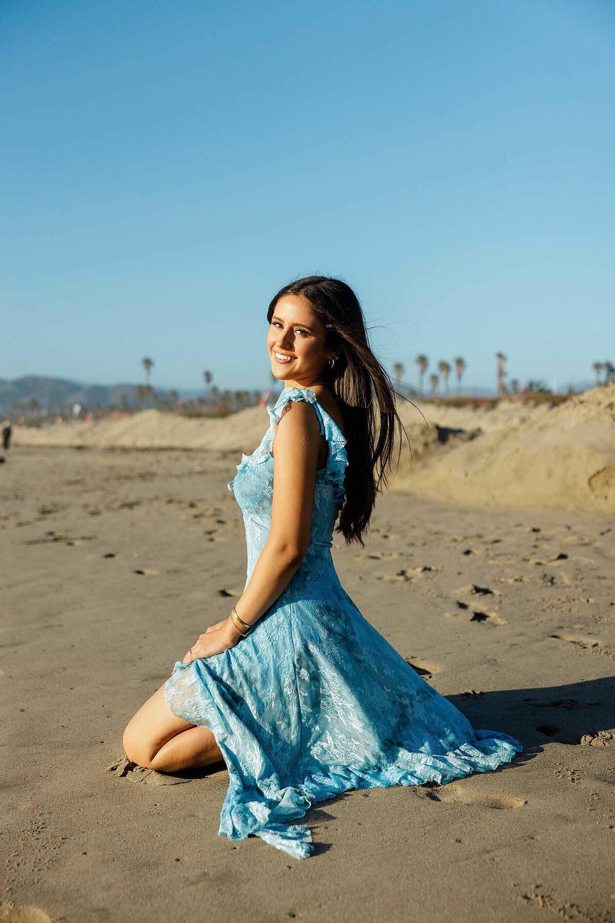 Senior girl in a blue dress smiling while kneeling in the sand at a beach near Los Angeles, with palm trees and mountains in the background