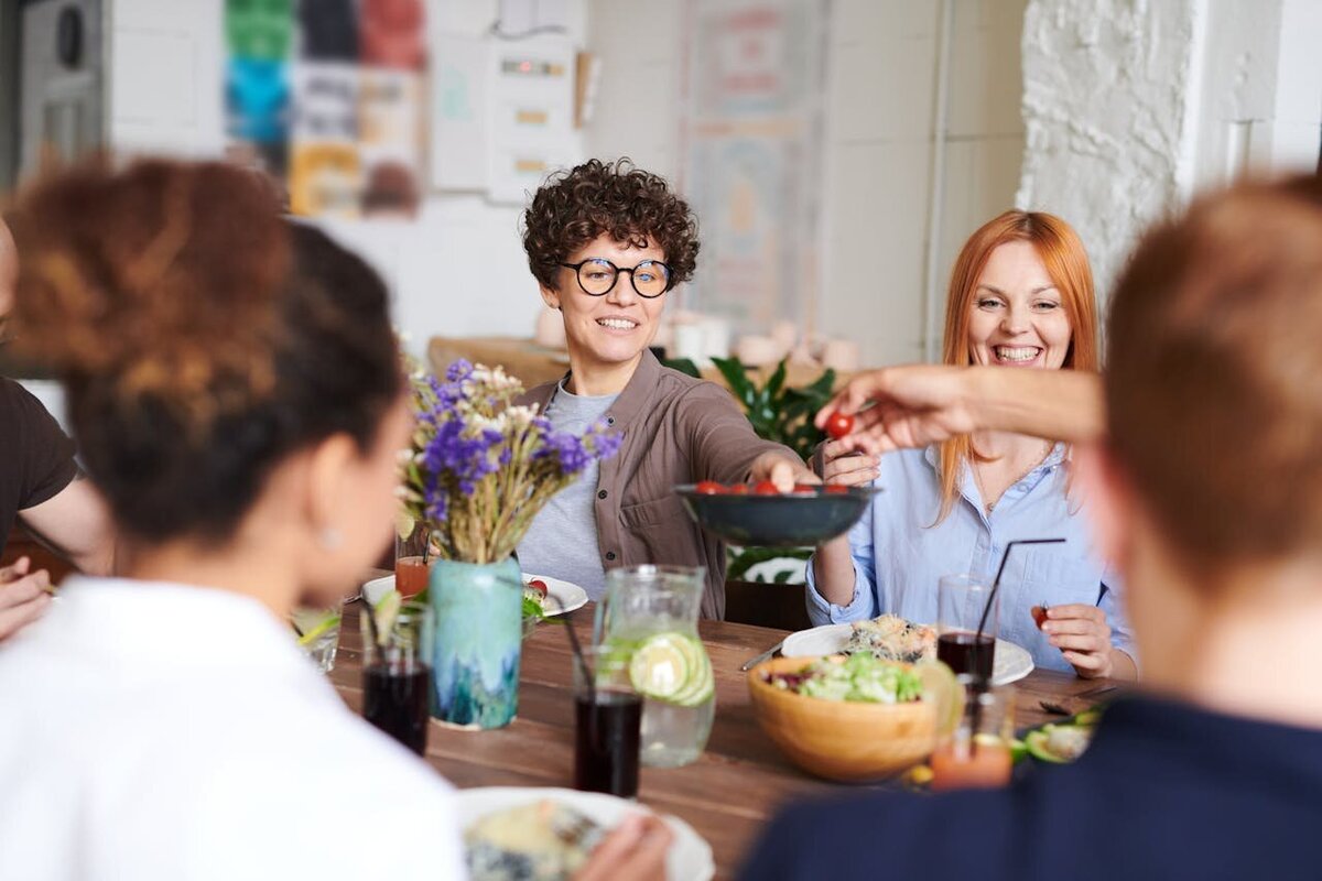 Group of people smiling and talking around a table with flowers representing connection and compassionate support for therapy at Rooted & Nourished Psychotherapy