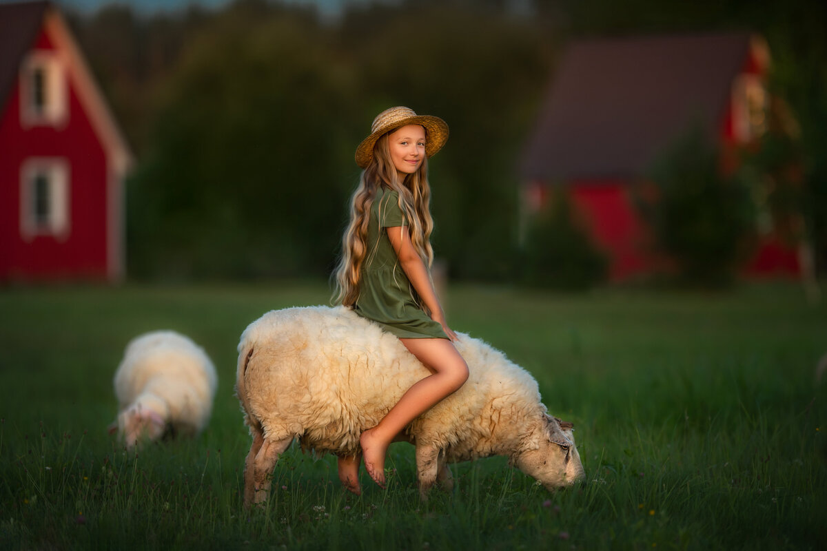 Amelia in green dress with long blond hair and straw hat is riding  the sheep in a village of Lithuania surrounded by tall grass and red little barn houses.
