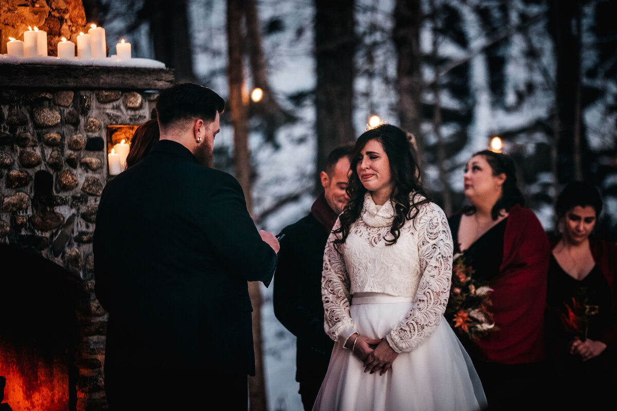 Bride looking at groom while he reads his vows in front of a warm fireplace in winter wedding at the Whitney's Inn in Jackson NH by Lisa Smith Photography