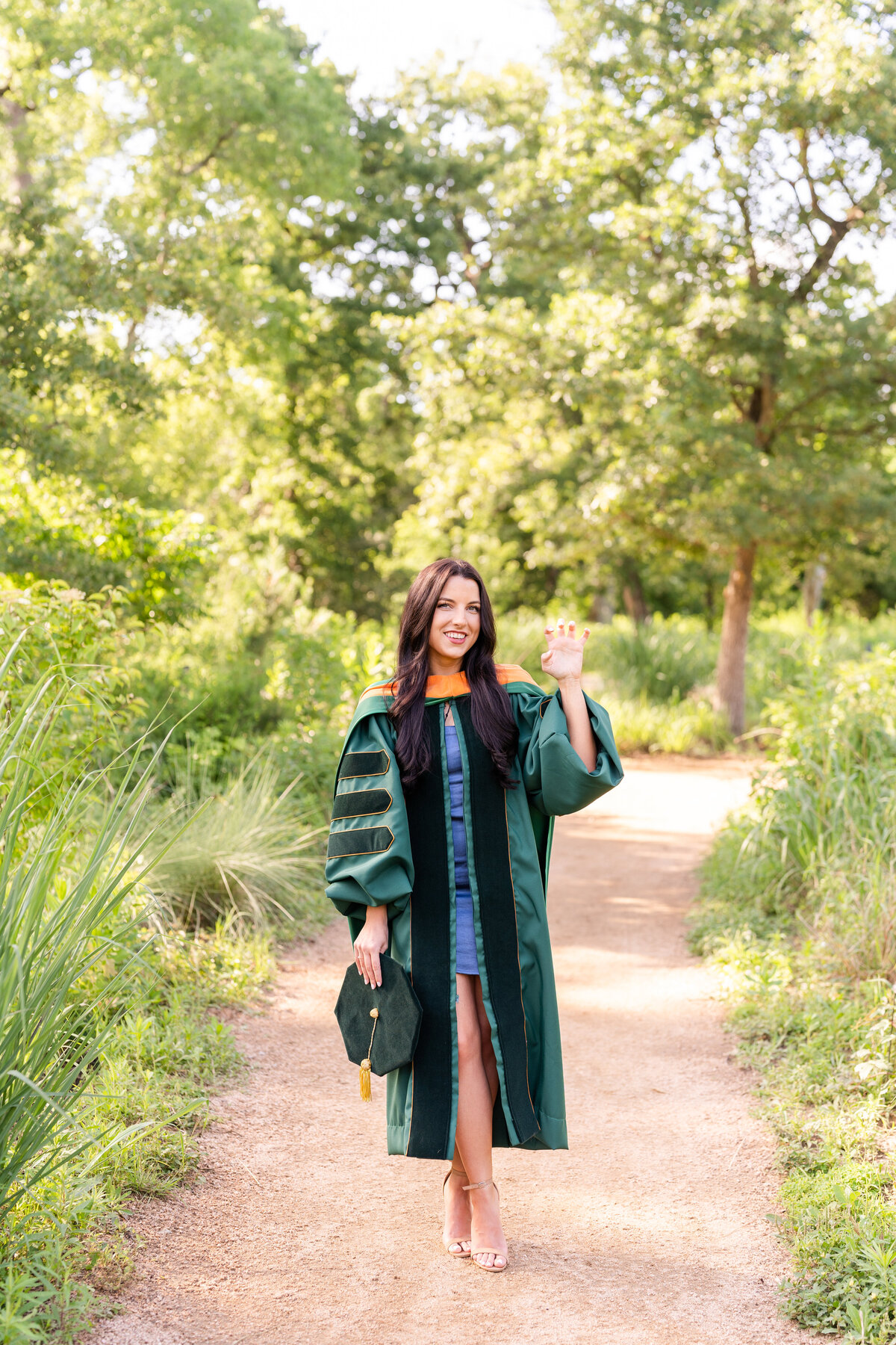 Baylor Doctoral Grad wearing gown, hood and holding hat while throwing up college hand sign and smiling at the Houston Arboretum
