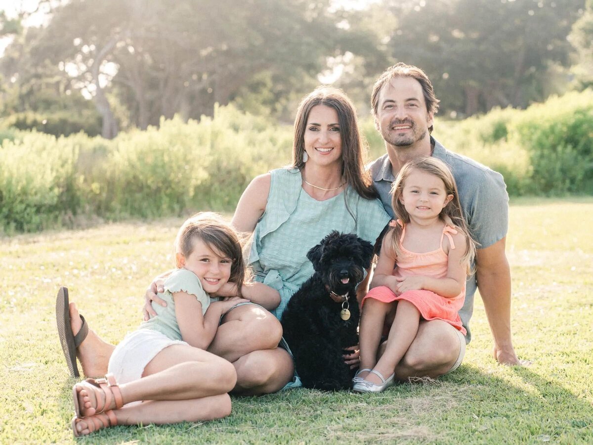 Parents sitting on the grass with two kids and a black dog.