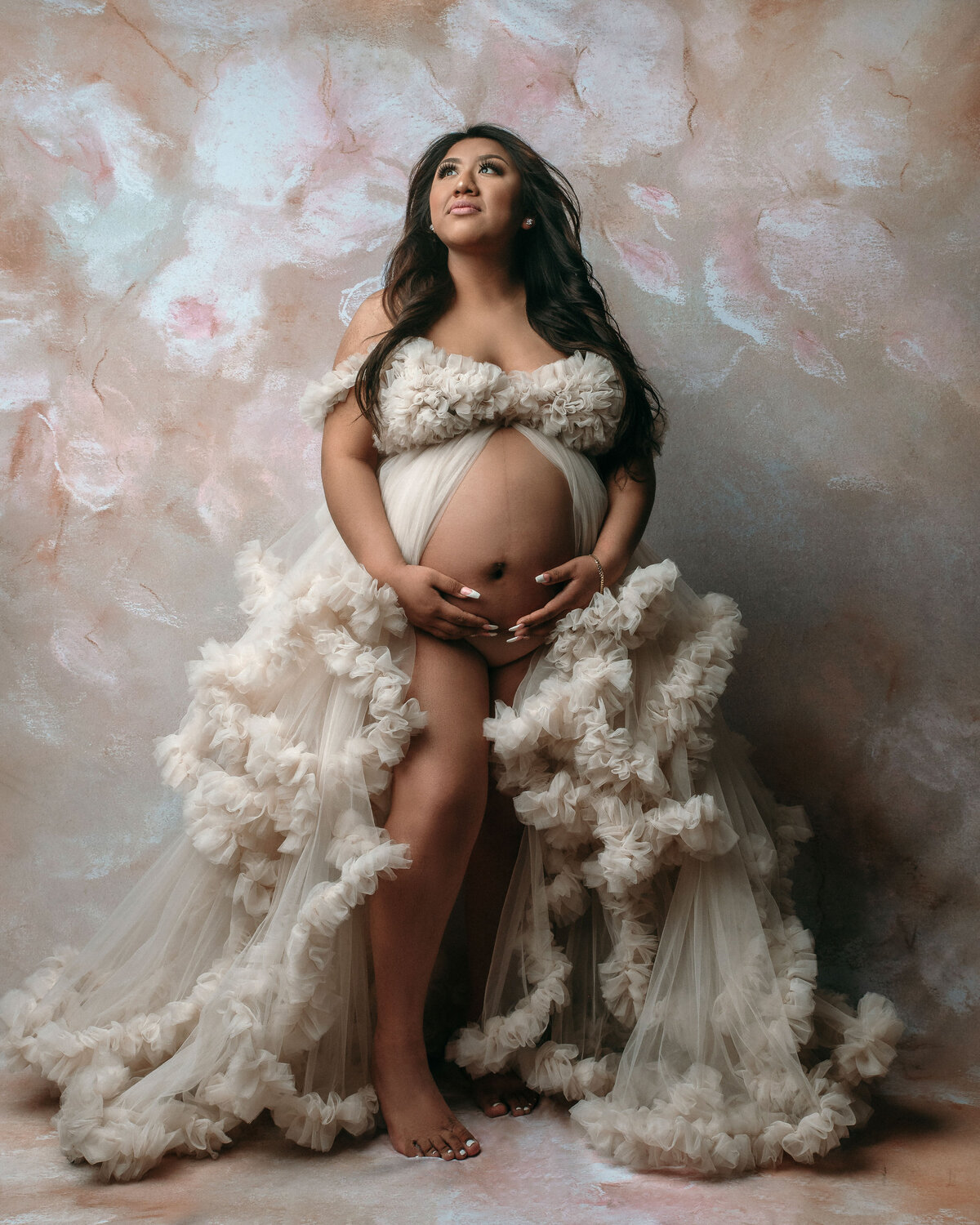 Pregnant woman looking up, holding tummy with both hands wearing cream tulle ruffle dress showing belly