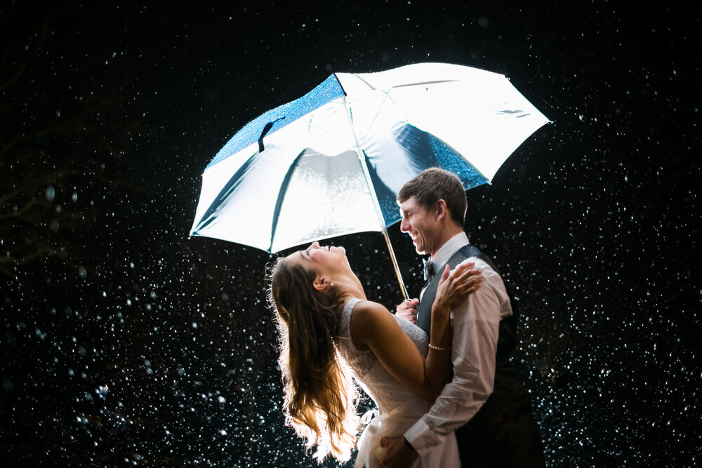 under an umbrella, groom grabs the bride's butt as she laughs during a rainstorm on their wedding day