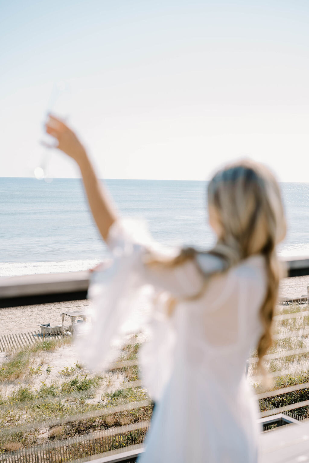 Rhode Island Wedding Photographers | A woman with long hair raises a glass toward a sunny beach, viewed from a balcony. The scene is bright and airy.