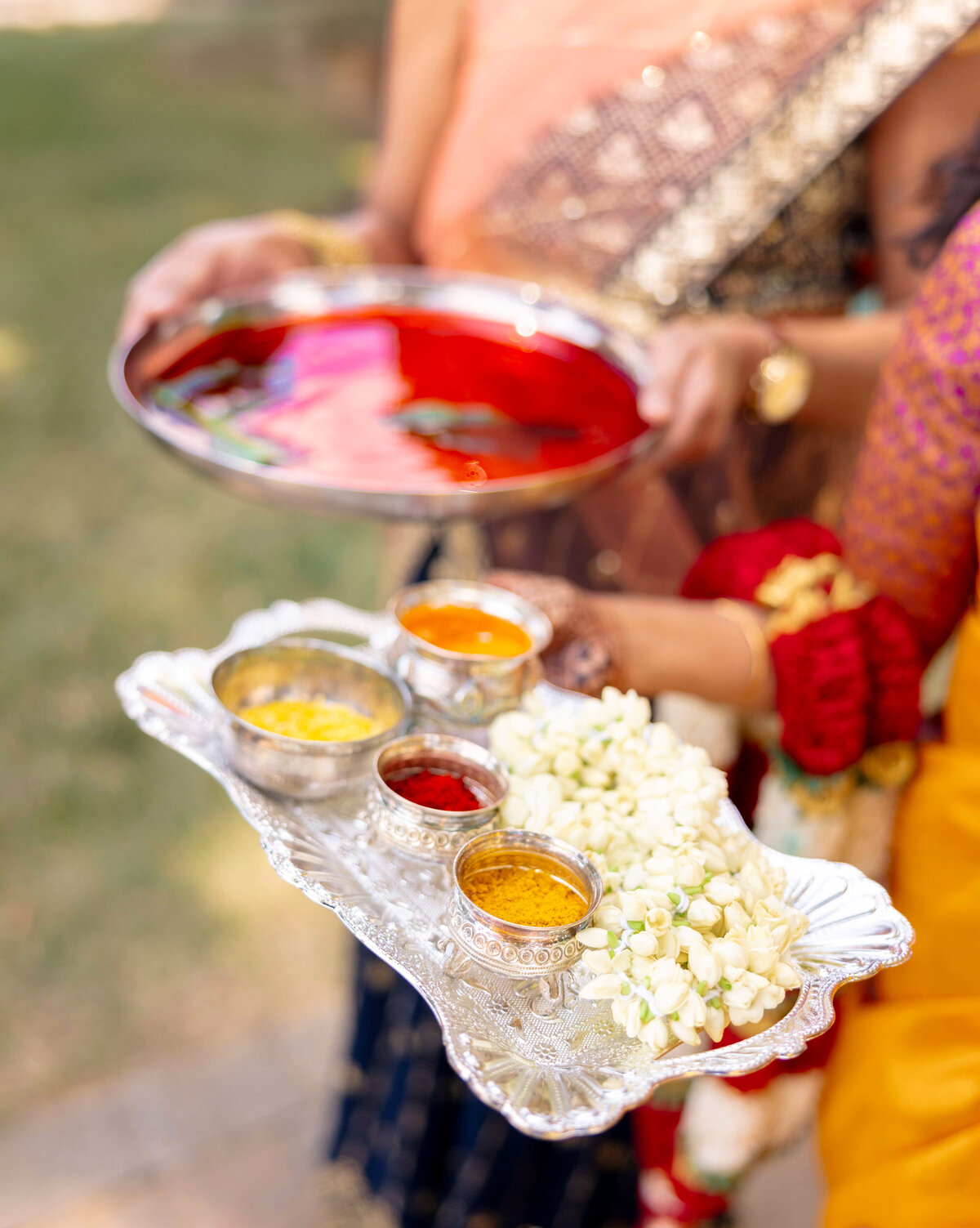 Close-up of two people holding silver trays during a traditional ceremony. One tray holds bowls of colorful powders and white flowers, while the other contains a red liquid with floating leaves. They are dressed in vibrant ethnic attire.