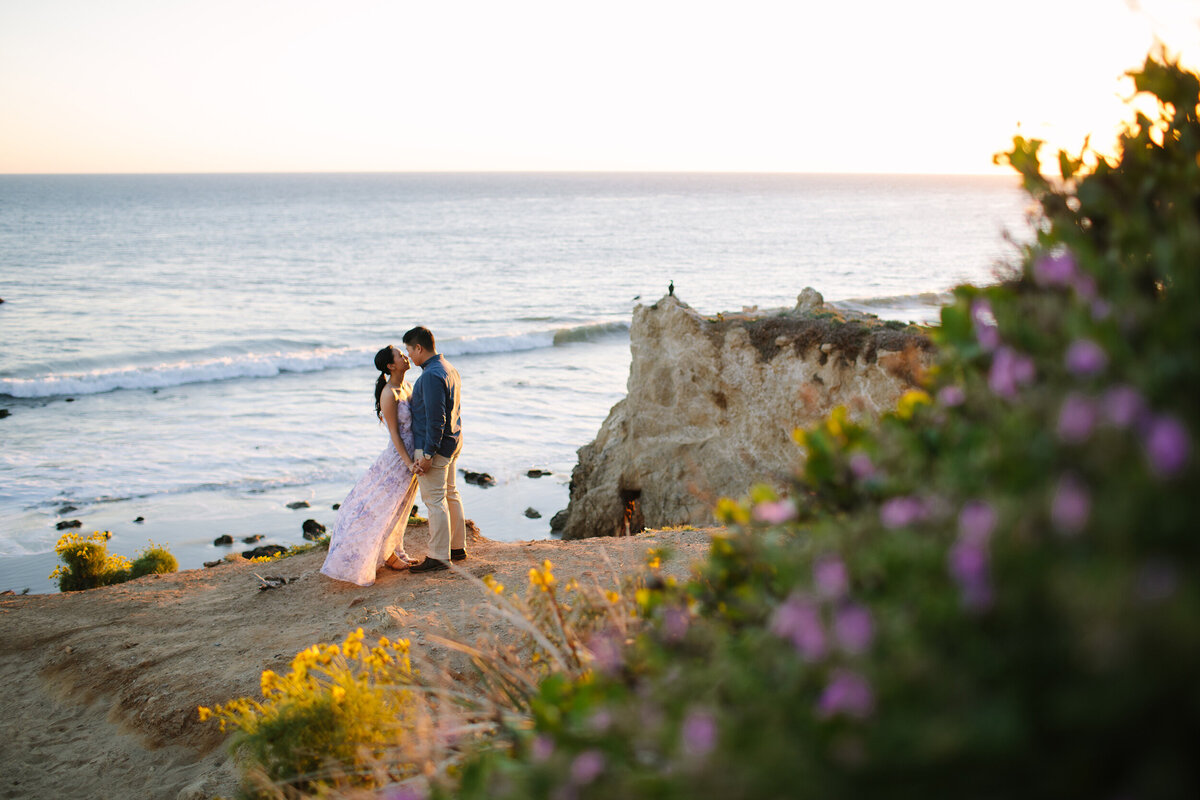 man and woman stand on the cliffside and kiss with the ocean in the background