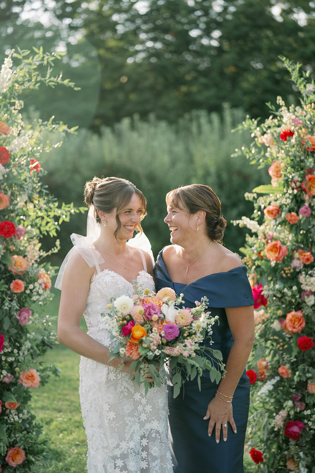 Bride and her mom standing and laughing together in front of the floral ceremony arch during their September orchard wedding at The Cherry Barn in Frankfort, Michigan.