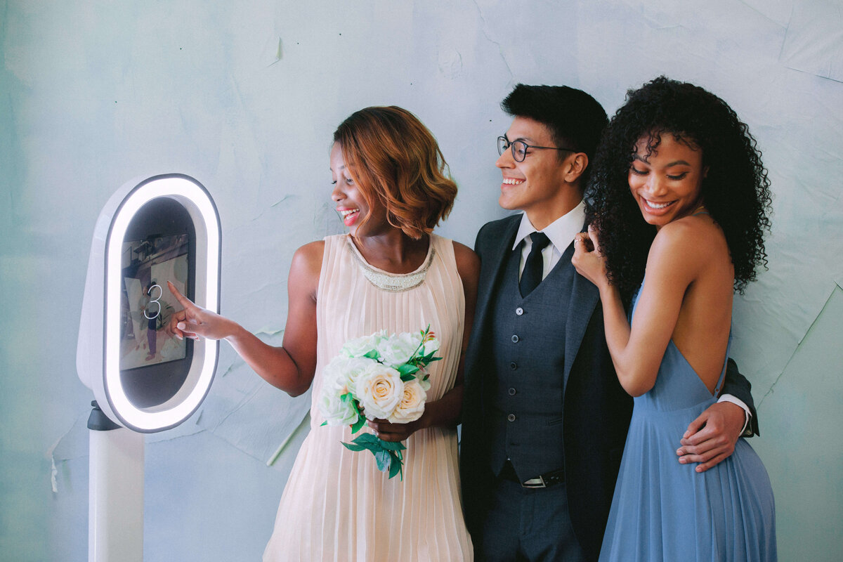 Lady wearing blue dress smiling, a man wearing suit and tie, and the other lady holding a bouquet touches the photo booth screen getting ready to have their photos taken