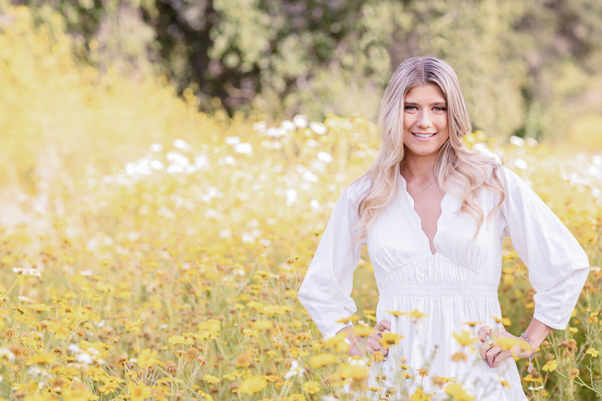 A woman with long blonde hair stands smiling in a field of yellow and white flowers. She is wearing a white dress with long sleeves. The background is filled with greenery and blossoms.