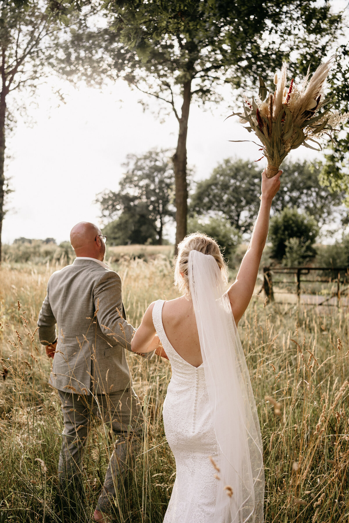 Golden hour shoot. Bruidspaar loopt door hoog gras en de bruid houdt het boeket omhoog. 