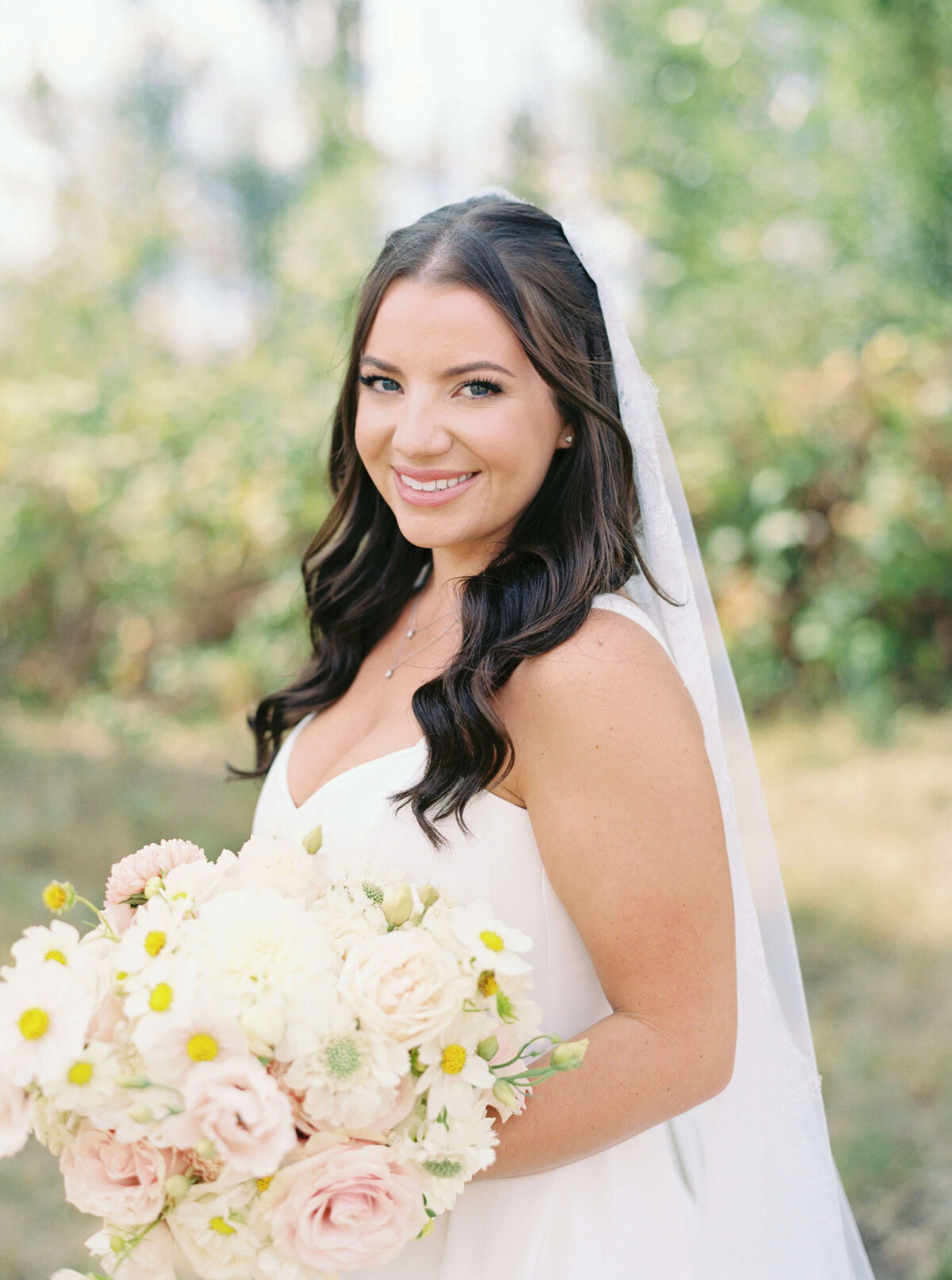 A bride wearing her wedding gown and veil smiles at the camera while holding her bouquet of flowers.