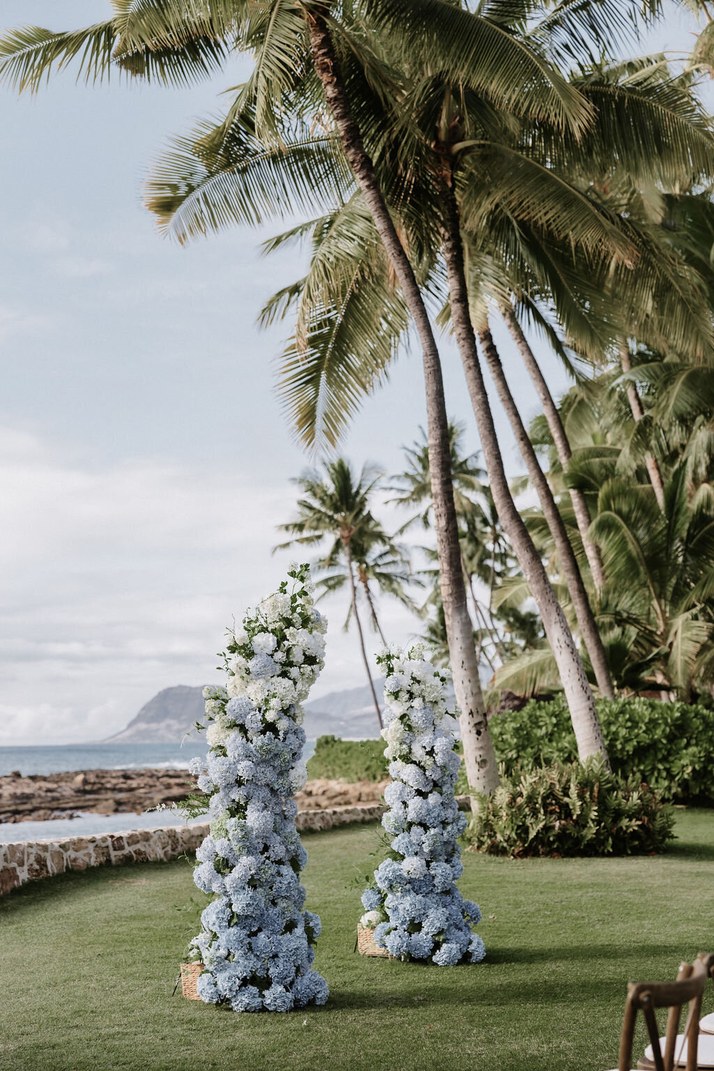 broken column wedding ceremony arch at lanikuhonua in front of the ocean with blue and white gradient hydrangeas and palm trees in the background