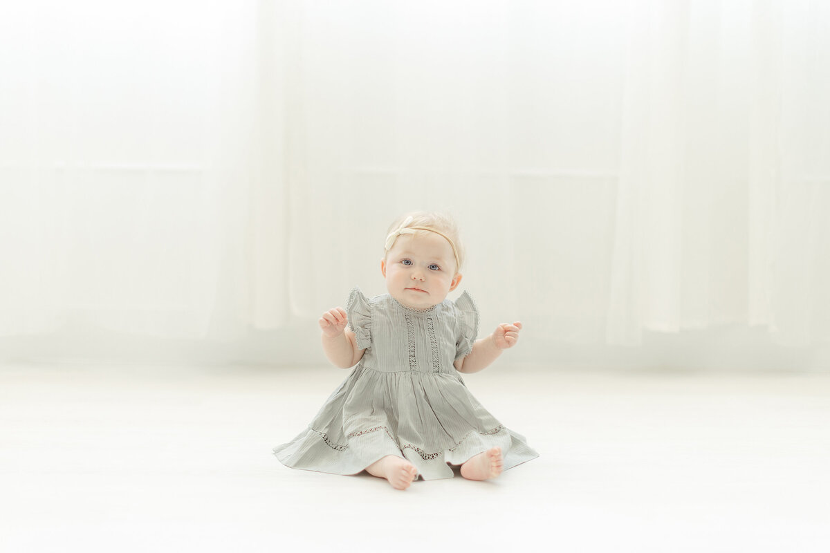 A baby girl sitting on a light filled studio floor looking at the milestone photographer as she captures her.