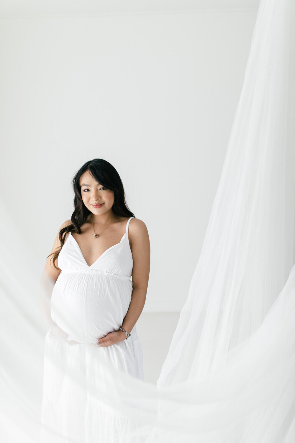 Asian woman who is pregnant stands next to white netting, wearing a white dress and looks at the camera