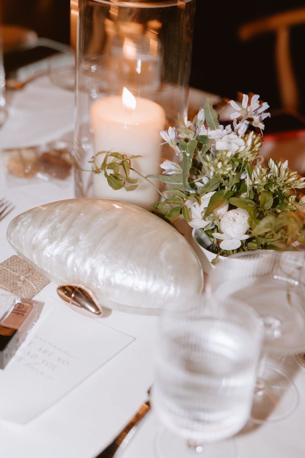Reception tablescape at night with pearl shell purse sitting on the table, a water glass, utensils, a candle and florals
