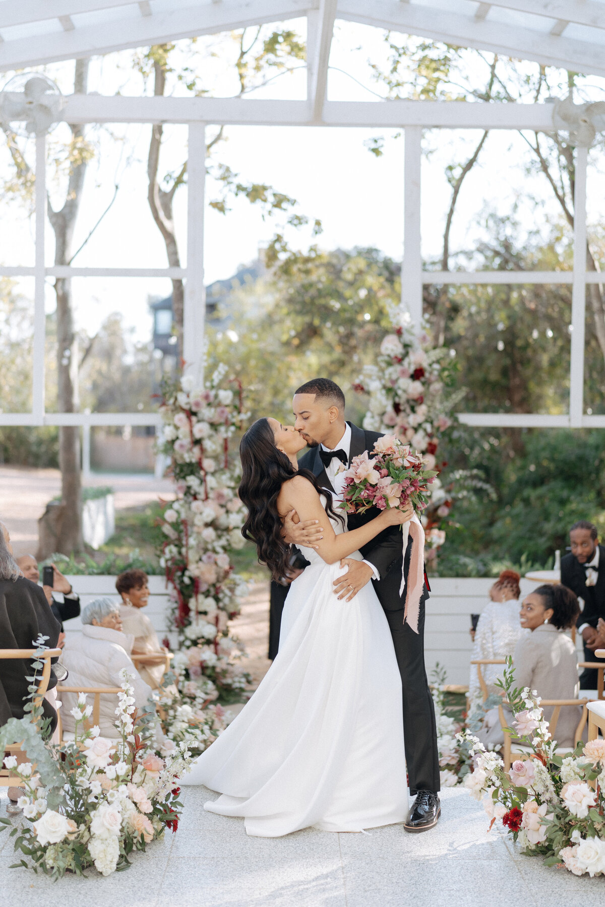 Bride and groom kissing beneath floral arches in a light-filled outdoor pavilion during a Houston wedding.