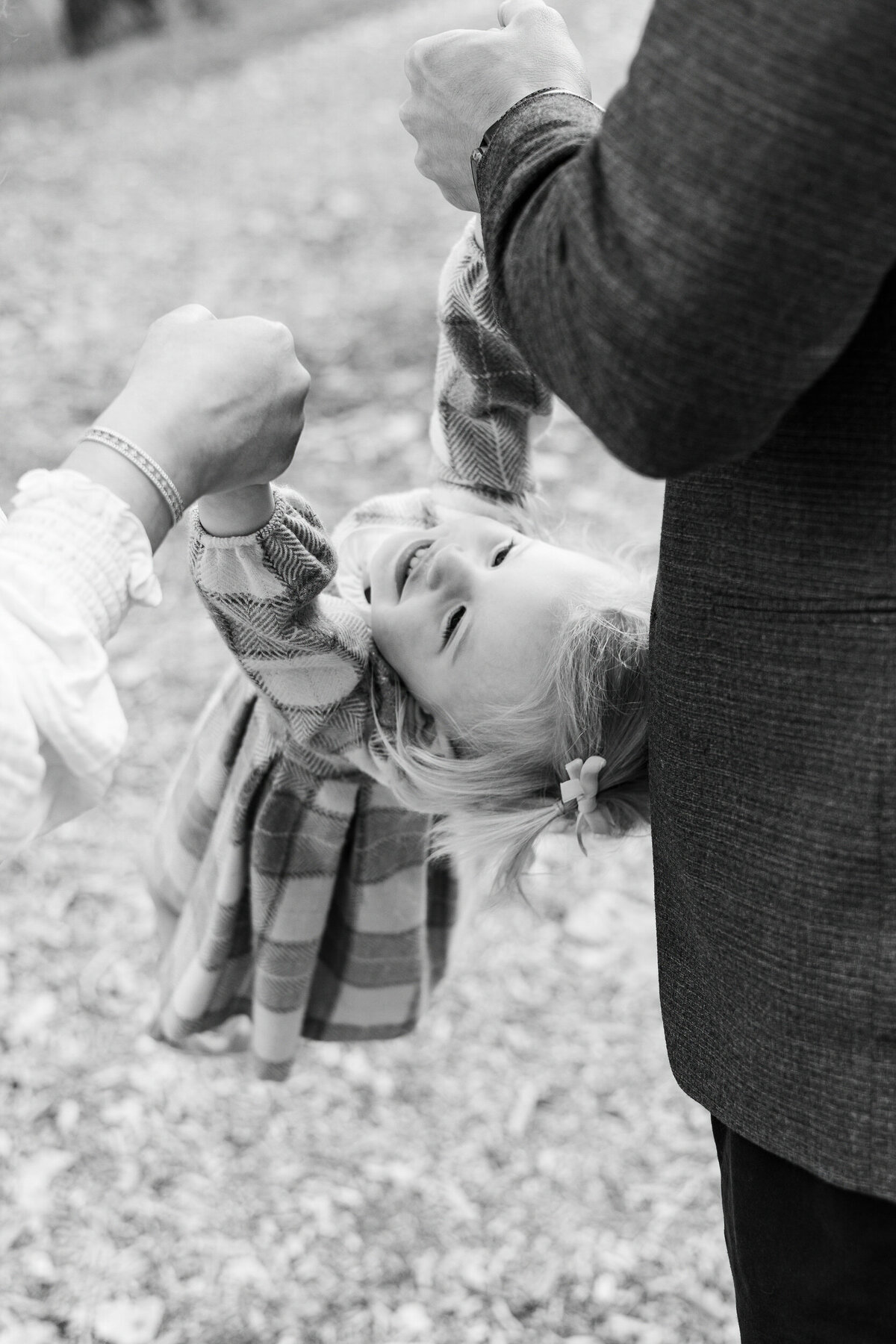 Black and white image of a toddler girl swinging from her parents' hands as she smiles.