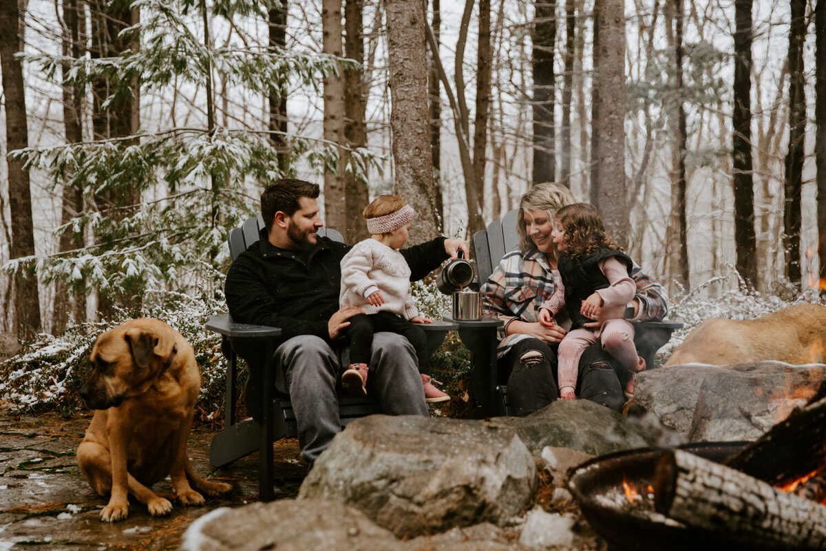 Mom, Dad, and two young daughters sitting amongst the trees at a styled outdoor campfire family photoshoot in London, Ontario. Dad is pouring hot chocolate and the family are smiling at each other. There is a campfire in the foreground.