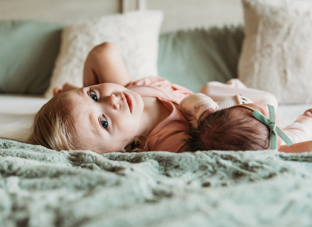 Newborn Photographer, Big sister looking at the camera while she's snuggling next to baby sister on the bed.