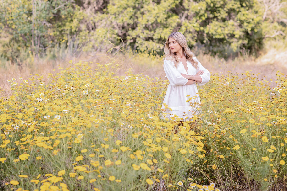 A woman in a white dress stands in a field of yellow wildflowers, with arms crossed, looking to the side. The background features green trees and a soft focus, creating a serene and natural atmosphere.