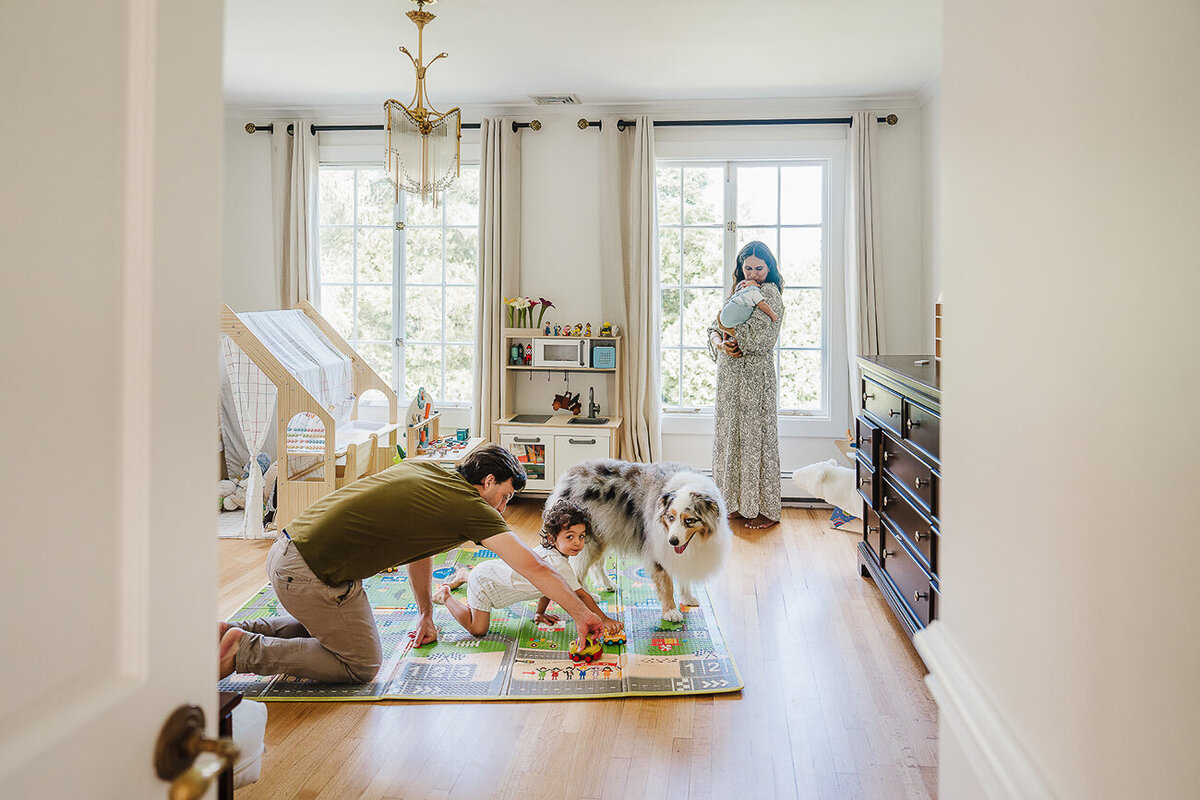 mother holds new baby in nursery while dad plays with toddler brother on floor
