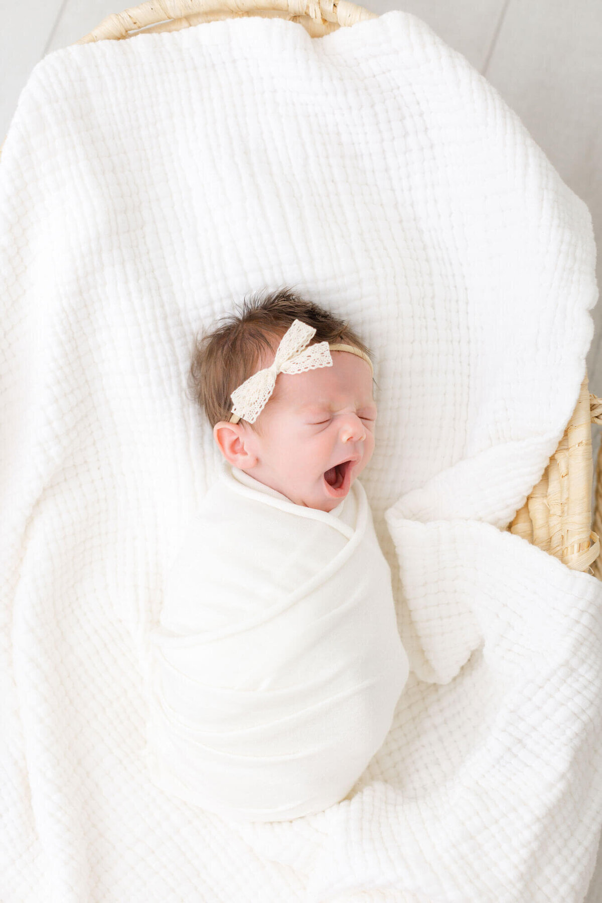 Baby swaddled in white and laying on a white blanket in a Moses basket. She is wearing a cream lace headband and yawning wildly.
