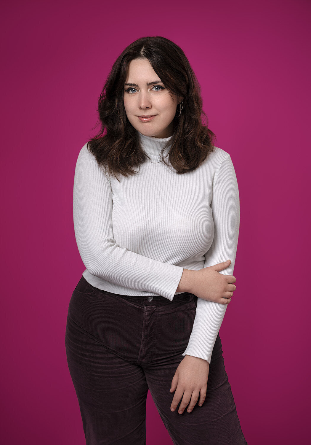 Image of a girl in a white roll neck top taken on a  bright pink background. She is holding her arm and smiling at the camera. Taken by Norwich Headshot photographer Claire Howes.
