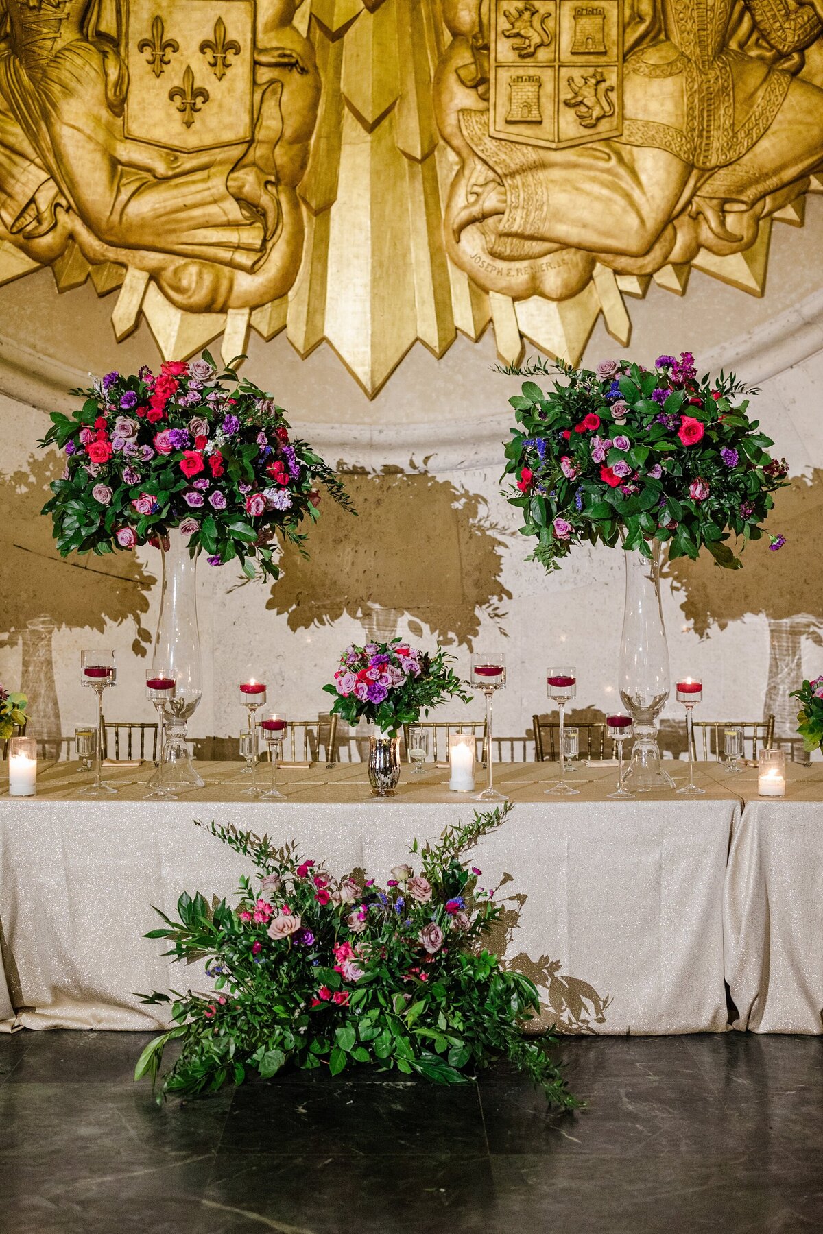 A detail shot of the head table at a wedding reception at the Hall of State at Fair Park in Dallas, Texas. The long, rectangular table is draped in with a tablecloth and covered with many candles and large floral arrangements in tall, glass vases. A large floral arrangement sits in front of the table on the ground and a large, gold crest adorns the wall behind the table.