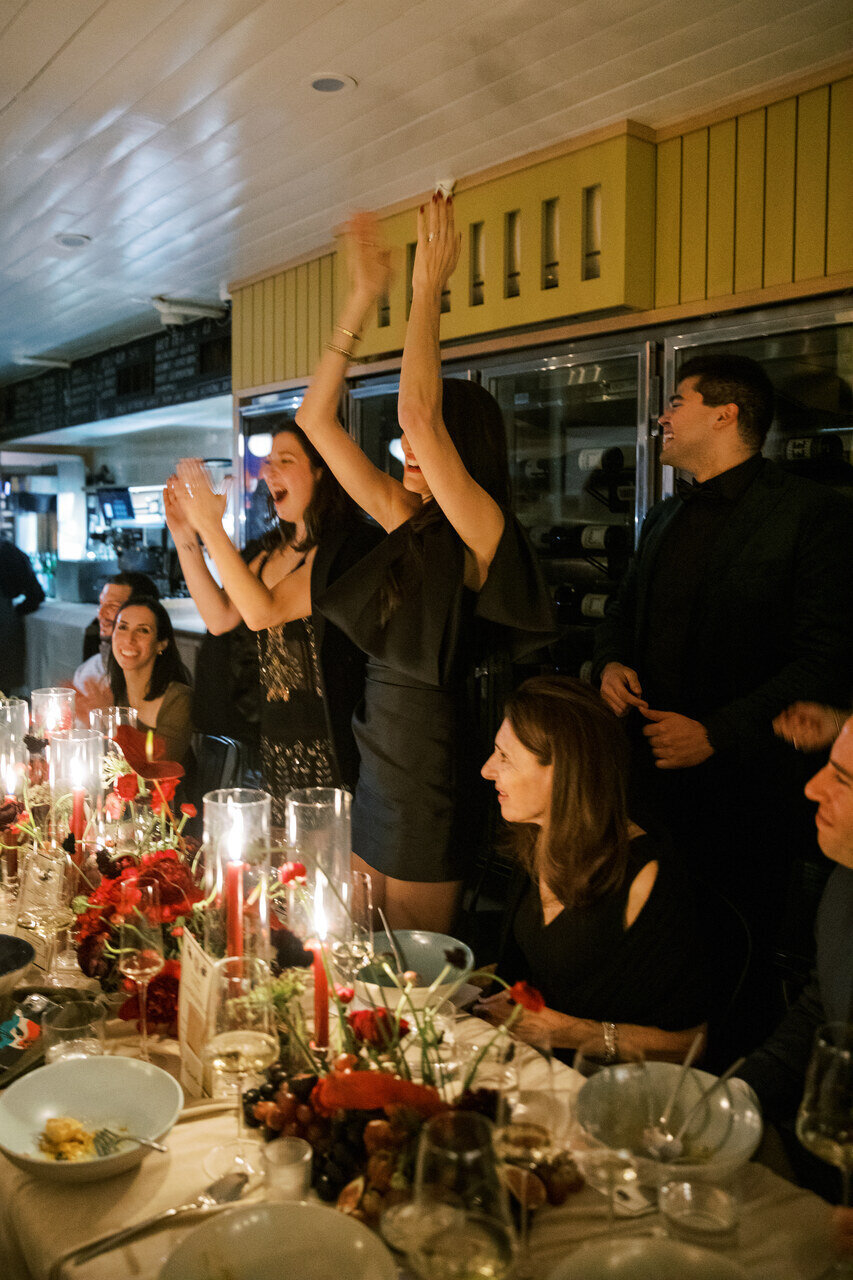 A lively dinner celebration with guests gathered around a beautifully set table with candlelight and floral arrangements. A woman in a black dress raises her hands in excitement as others smile and engage in conversation.