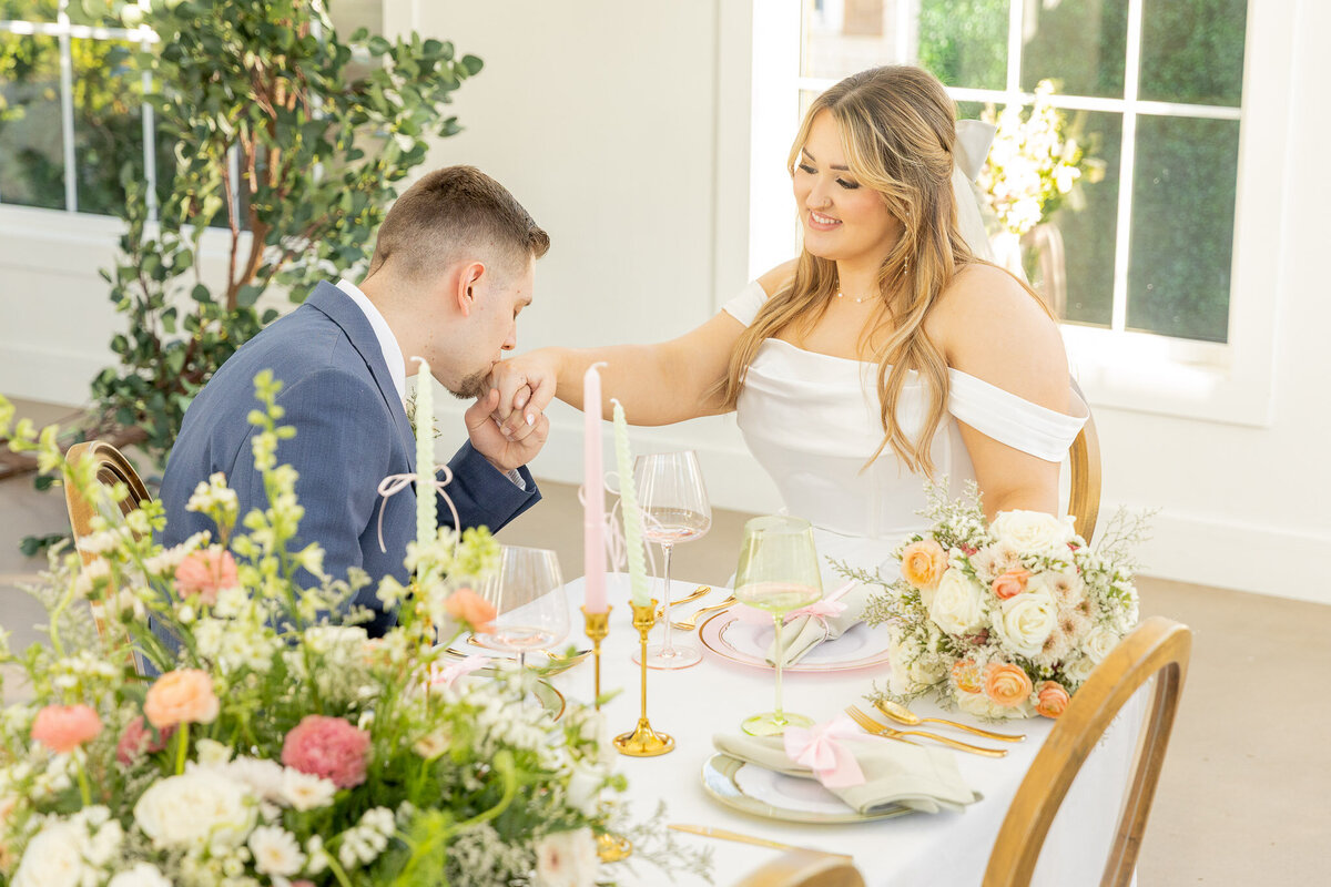 A groom kisses the hand of his bride on their wedding day. She's sitting in a chair and he's kneeling down so he's on her same level.