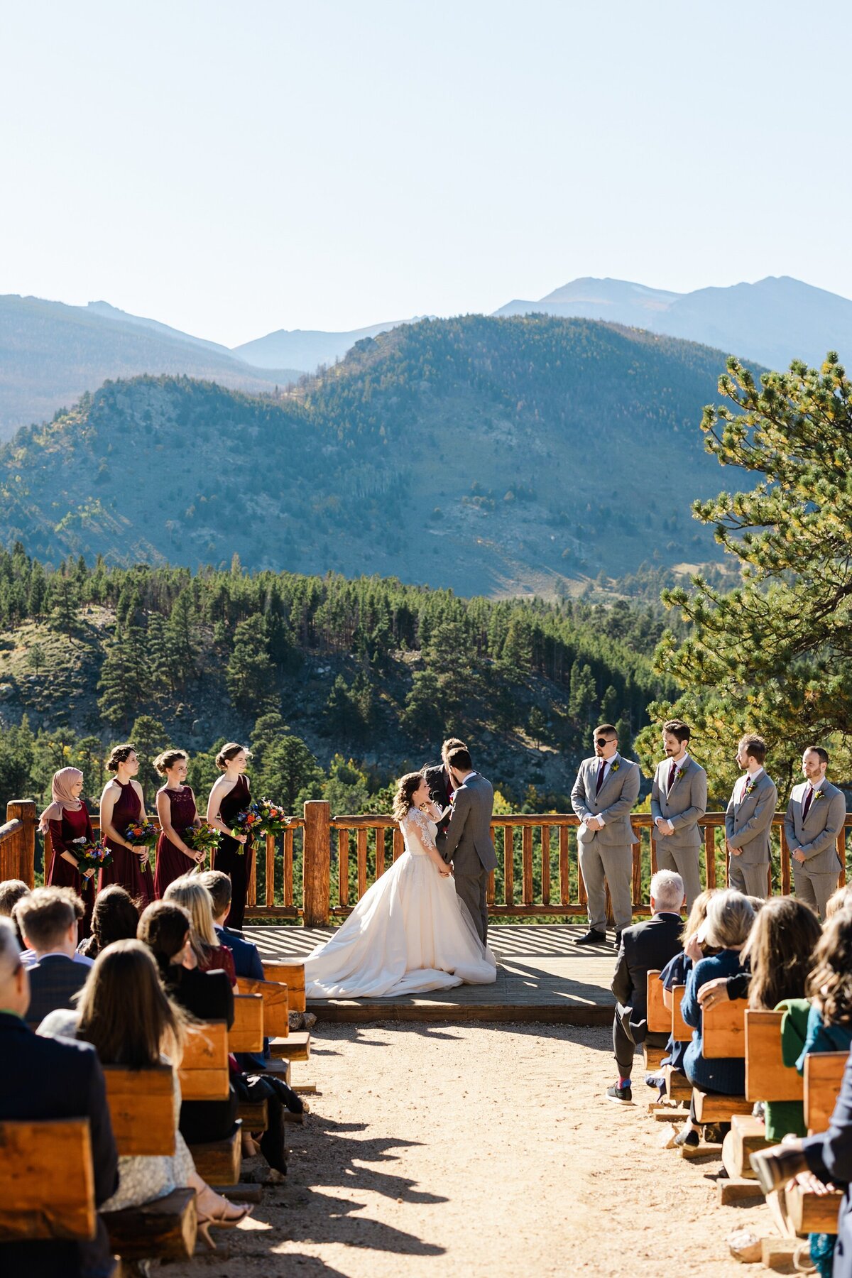 Candid shot of a bride and groom during their wedding ceremony at the YMCA of the Rockies in Estes Park, Colorado. The couple face towards each other in front of their officiant while their wedding party flanks them on either side. Many guests can be seen in the foreground while rows and rows of trees and mountains can be seen off in the distance behind the couple.