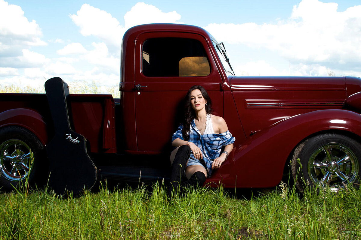 Country Musician Photography Portrait Female Artist Kristin Carter sitting in grass field infront of vintage red truck black guitar case leaning beside her