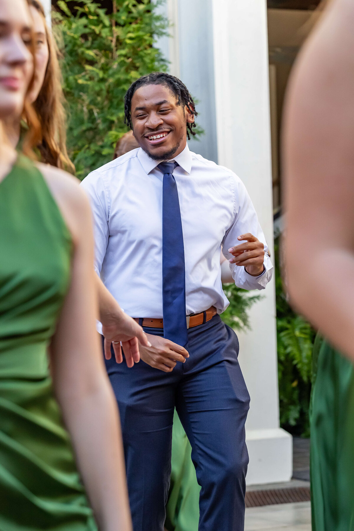 A man in a white shirt and blue tie is smiling and dancing outdoors, surrounded by people wearing green dresses. Lush greenery is visible in the background.