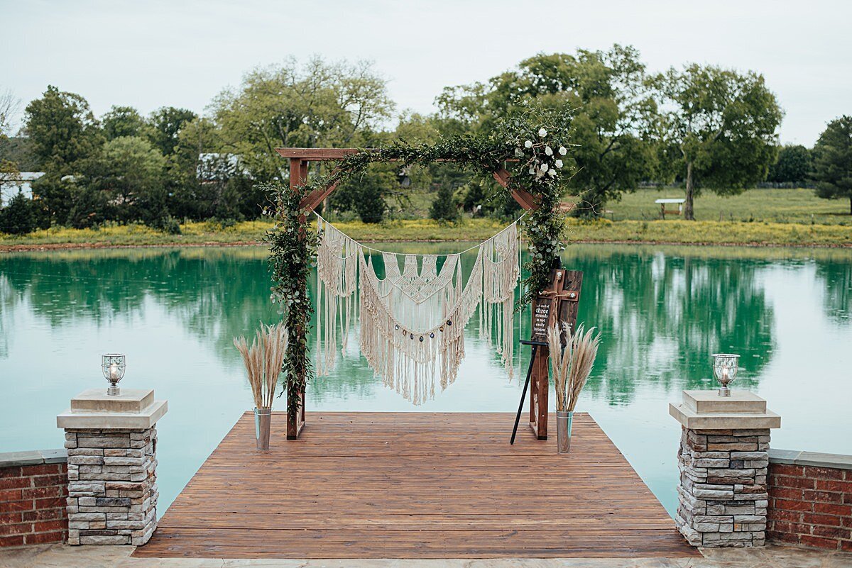 Lakeside wedding ceremony at Steel Magnolia Barn with a wooden arbor decorated with greenery, white and burgundy flowers and a boho macrame arbor hanging with tall vases of pampas grass at Steel Magnolia Barn.