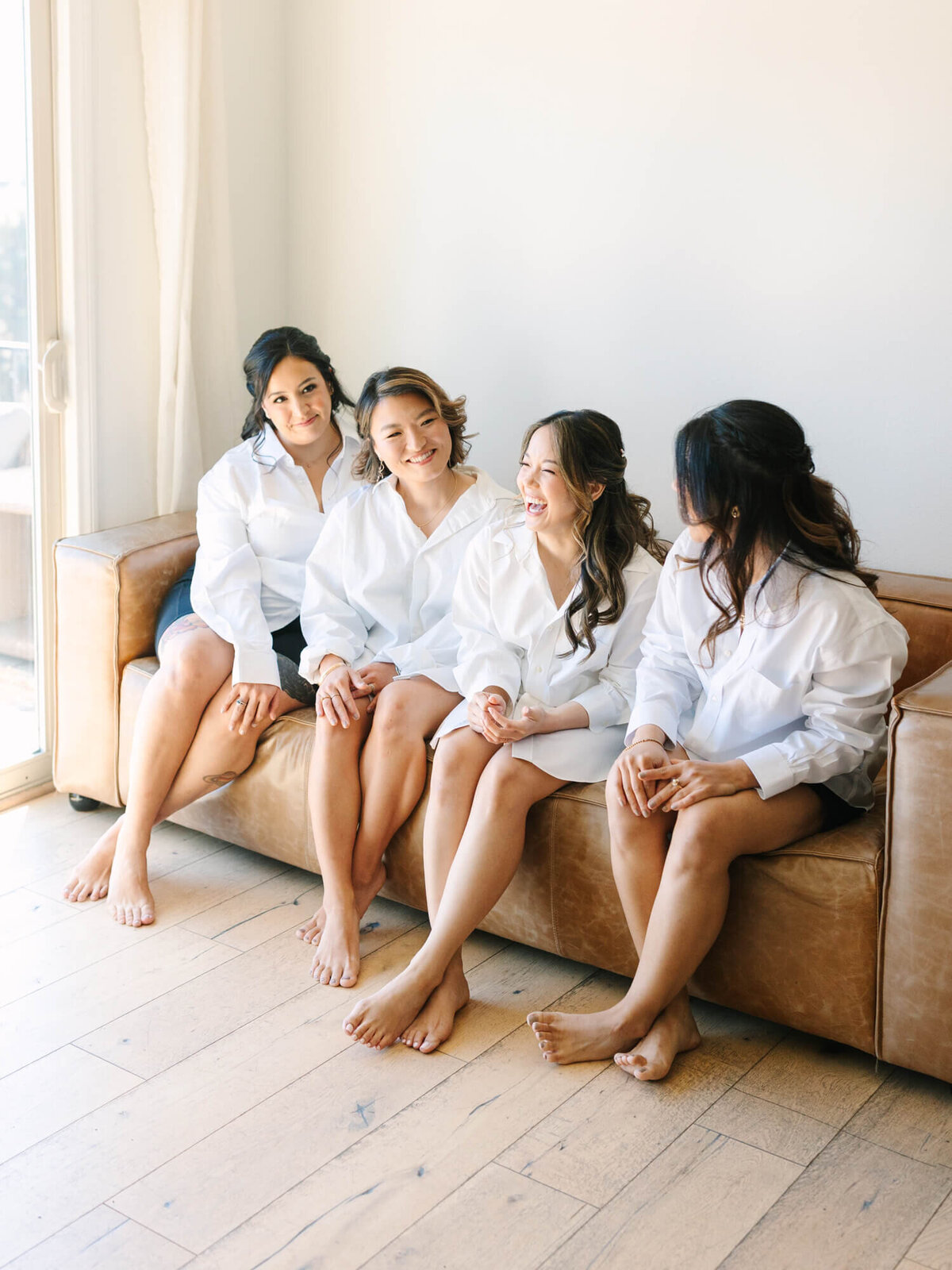 Bride and three bridesmaids sit on a leather sofa, wearing white shirts, smiling and chatting.