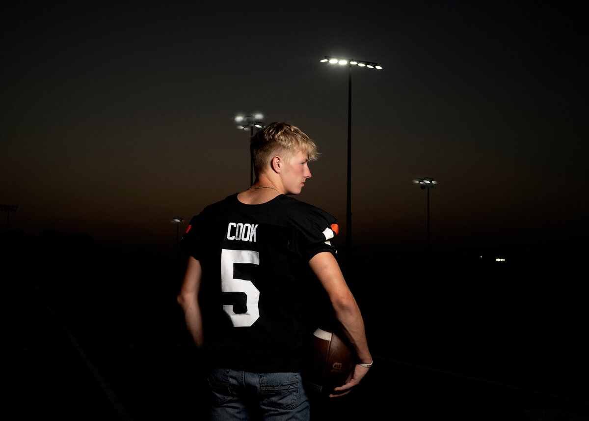 A football player in high school stands by the stadium at night with lights on in his jersey and holding a football