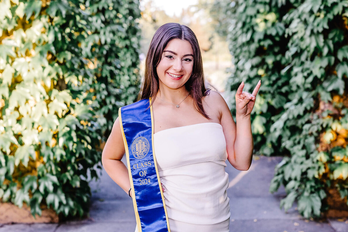 A woman in a white dress stands on a path bordered by green foliage. Shes smiling, wearing a blue graduation stole, and making a hand gesture with two fingers extended up.