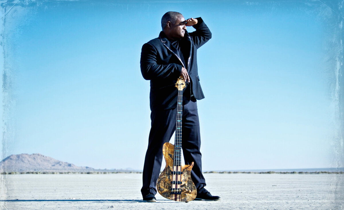 Male musician portrait Mitchell Coleman Jr wearing black suit holding bass guitar between legs hand across forehead looking out amidst desert background