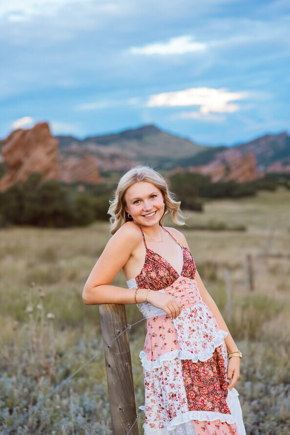 Girl in patchwork dress leans on post for her senior photo session.